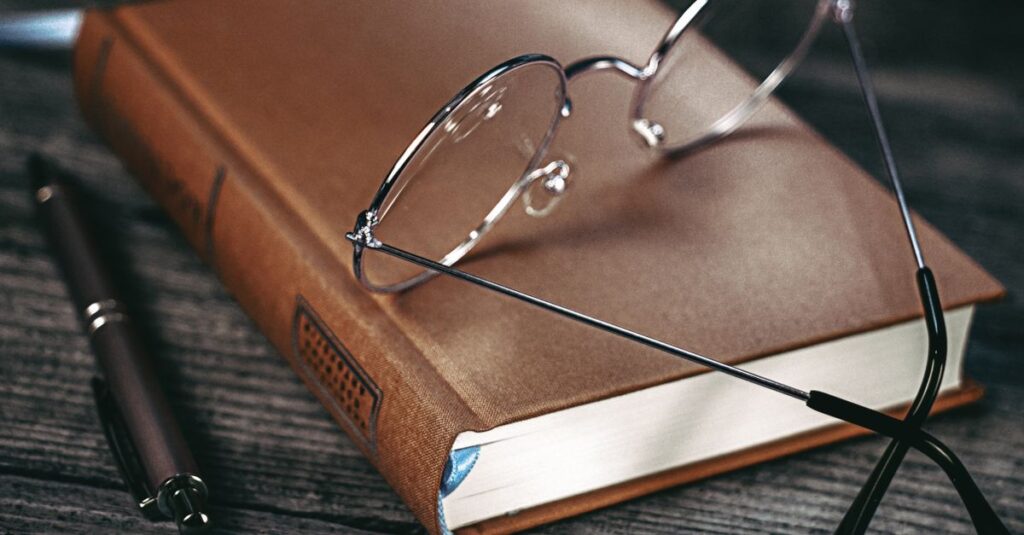 A brown hardcover book with eyeglasses and a pen on a wooden table.